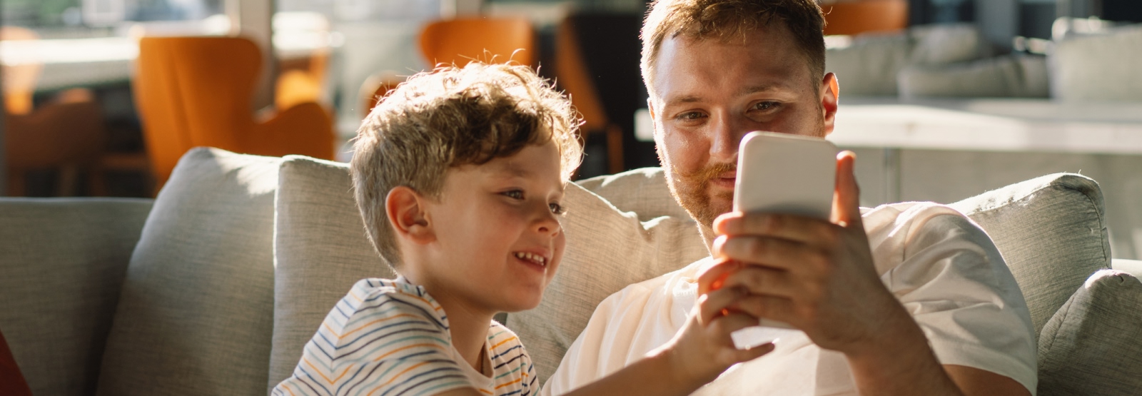A dad and young kid sitting on a couch and looking at a smartphone